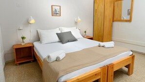 Cama bem arrumada com travesseiros brancos e cinzas, um cobertor bege e duas toalhas enroladas. Há duas mesas de cabeceira com abajures e uma pequena planta. Um guarda-roupa de madeira e um espelho estão na parede.