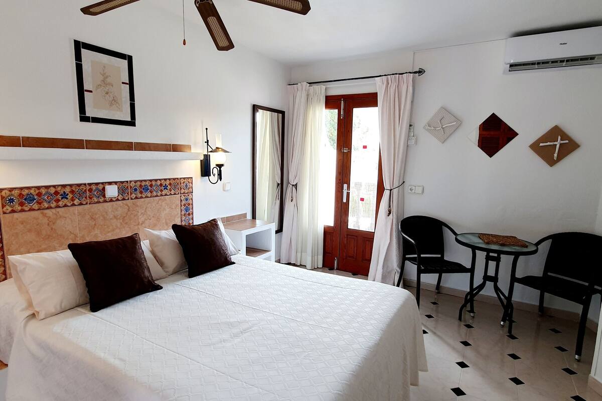 Une chambre d’hôtel comprenant un lit, une table de chevet, une lampe fixée au mur, une tête de lit décorative et une petite table avec des chaises.