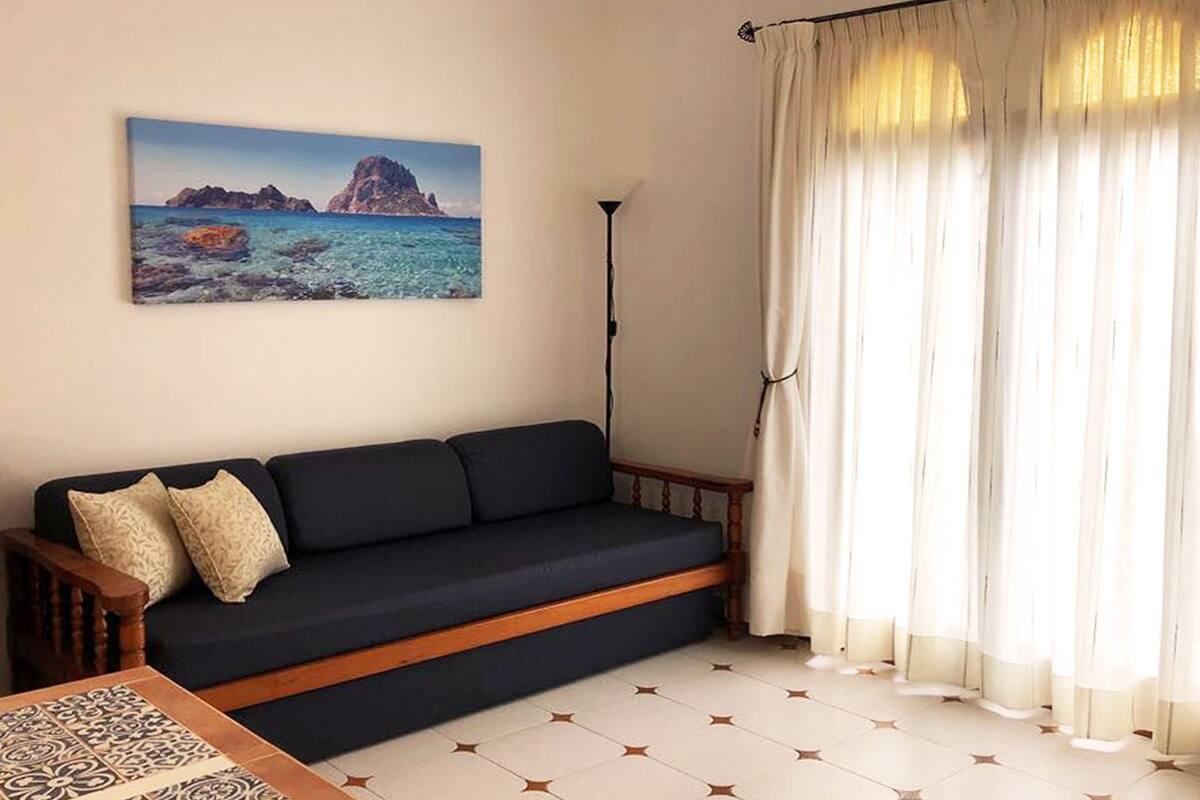 Une pièce avec un canapé bleu foncé, un tableau représentant un paysage côtier, un sol recouvert de carreaux à motifs étoilés et de fines rideaux.