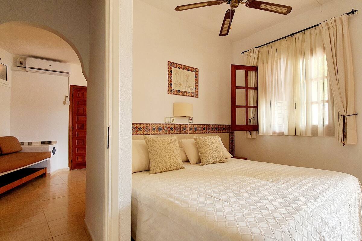 Une chambre à coucher avec un lit, un ventilateur de plafond, une fenêtre avec des rideaux et une petite table près de la fenêtre.