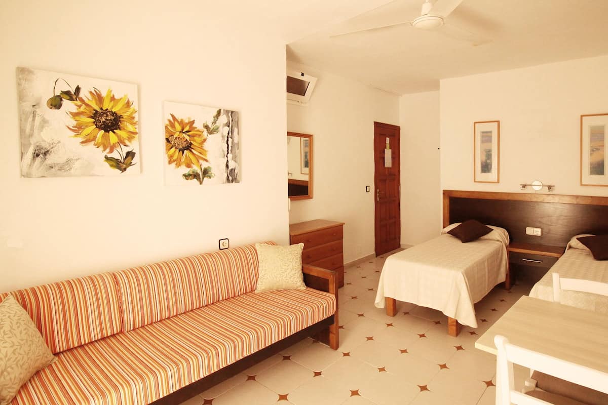 Une chambre d’hôtel avec un canapé à rayures, deux lits simples, une commode en bois et deux tableaux représentant des tournesols, accrochés au mur.