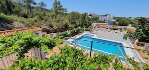 A pool area with a view of a hillside, surrounded by greenery and a building in the background.