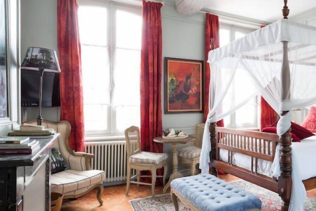 A four-poster canopy bed with red pillows, a bedside table, a lamp, and a chair in a room with a window and a radiator.