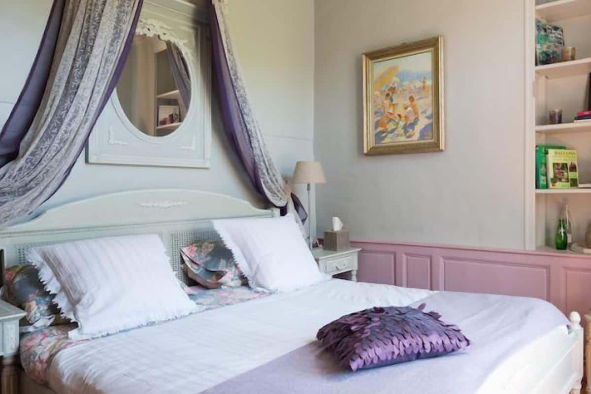 A bedroom with a four-poster bed, a nightstand with a lamp, a chair, and a bookshelf with books and decorative items.