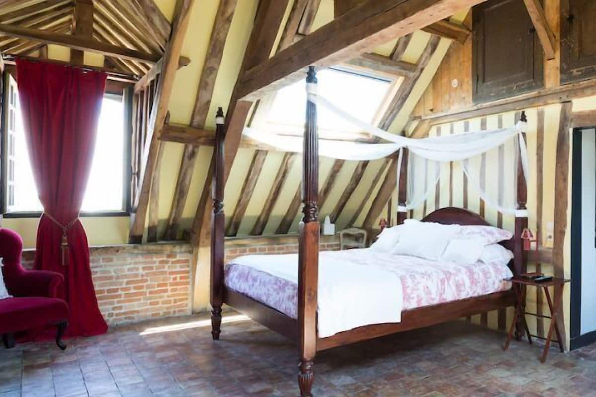A bedroom with a four-poster bed, a red armchair, a white chair, and a wooden cabinet.