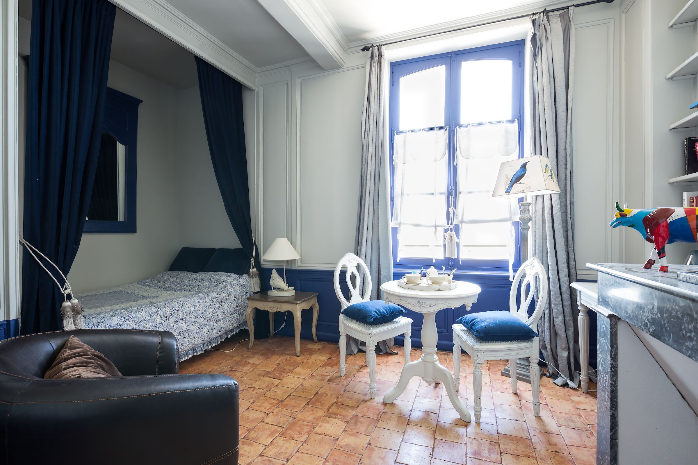 Una habitación con cama, escritorio, dos sillas, lámpara y una mesita con un objeto decorativo.