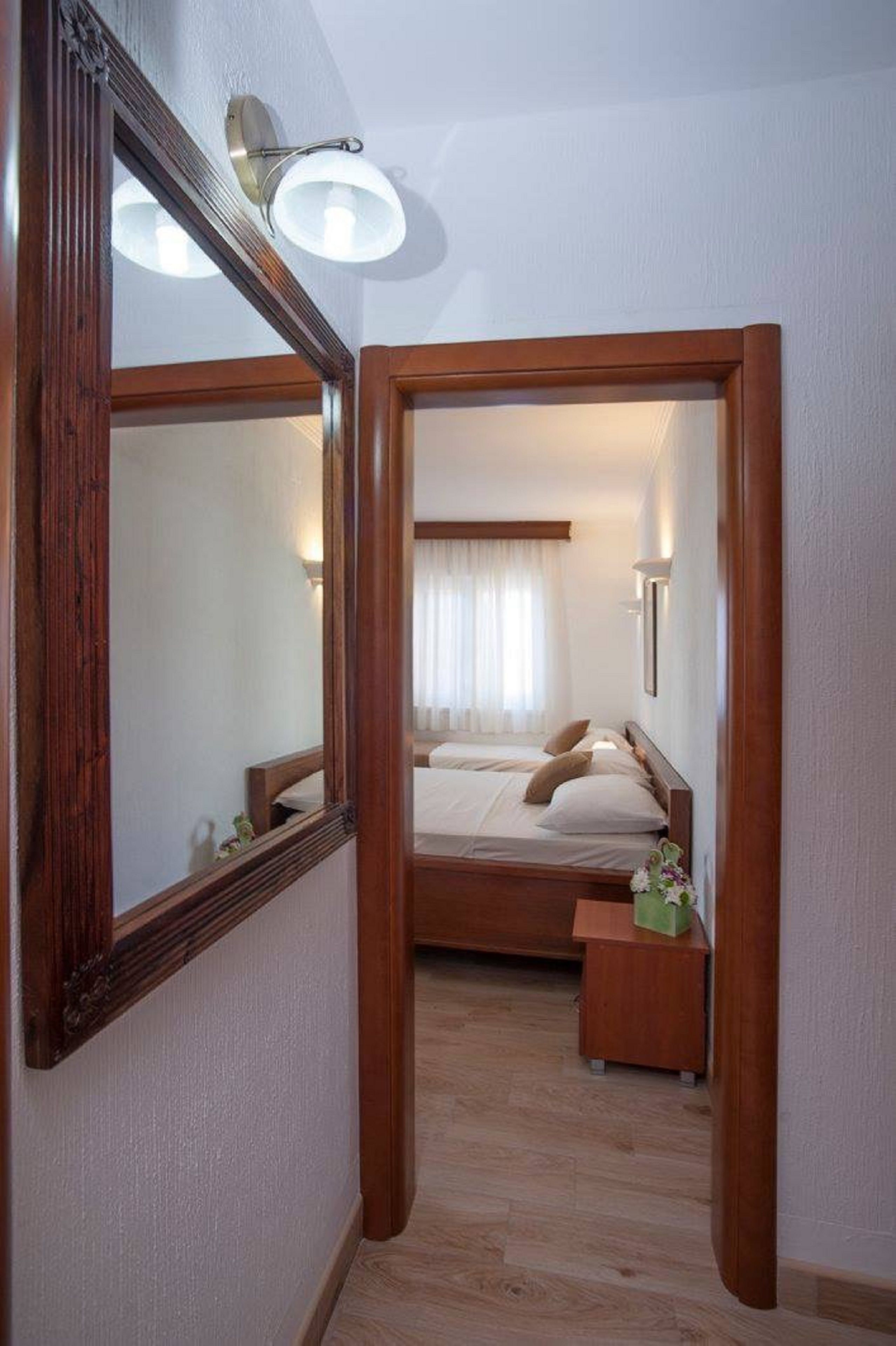 A bedroom with a large mirror, a bed with white linens, a nightstand, and a small table with a vase.