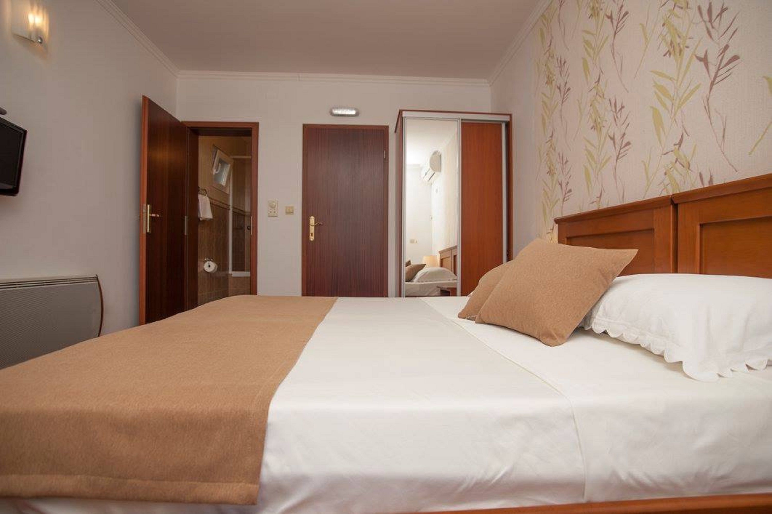 Ein Hotelzimmer mit einem Bett, einem hölzernen Kopfteil, einer weißen Wand, einem an der Wand befestigten Fernseher und einer Tür zu einem Badezimmer.