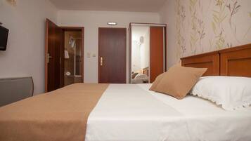 Ein Hotelzimmer mit einem Bett, einem hölzernen Kopfteil, einer weißen Wand, einem an der Wand befestigten Fernseher und einer Tür zu einem Badezimmer.