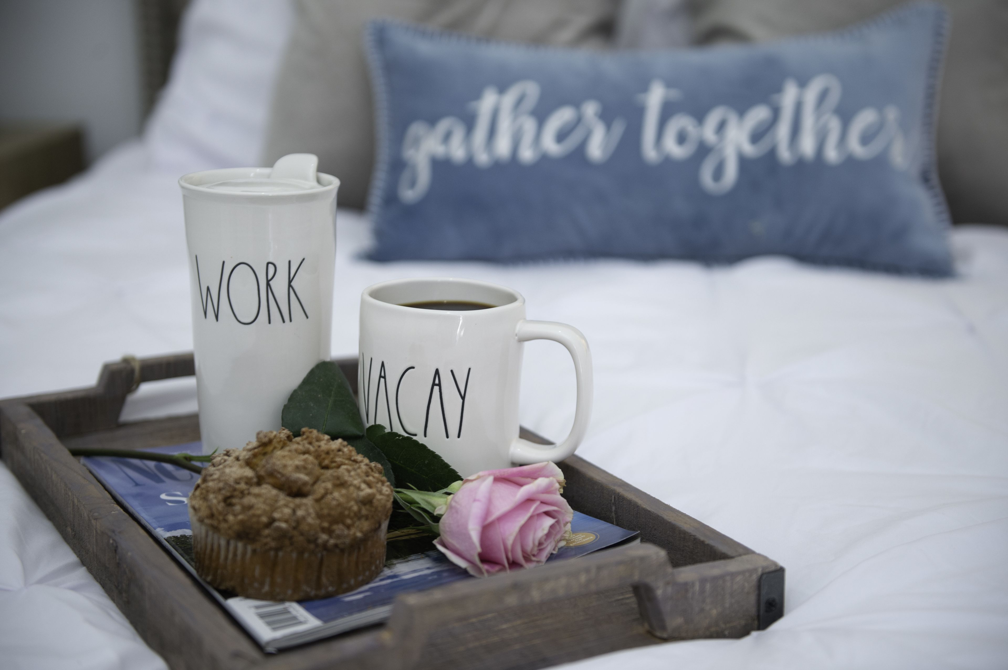 Una bandeja con un muffin, una taza de café con la etiqueta 'WORK' y otra taza de café con la etiqueta 'VACAY' sobre una cama con un almohadón que dice 'gather together'.