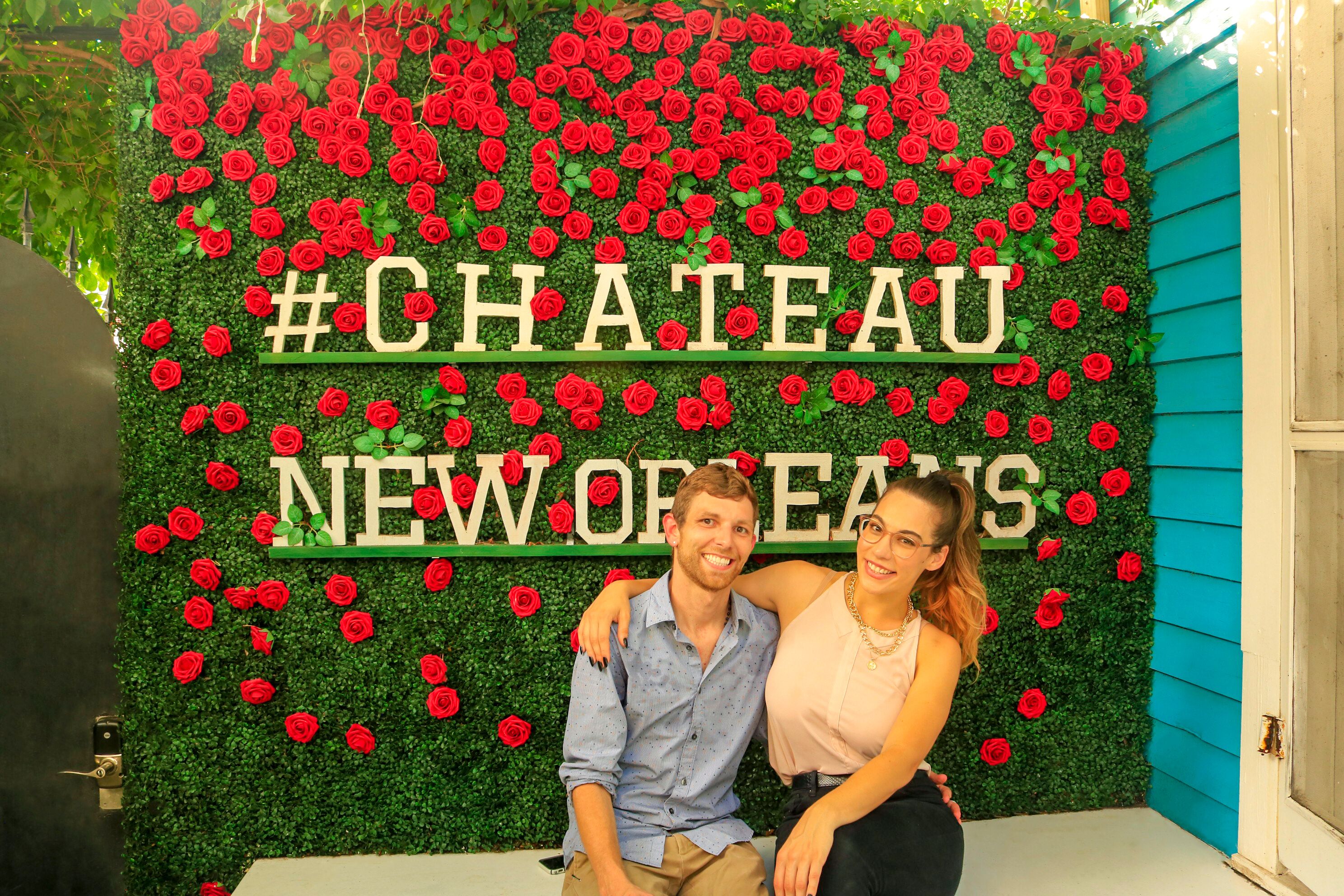 Una pareja sentada frente a una pared decorada con rosas rojas y el texto '#CHATEAU NEW ORLEANS'.