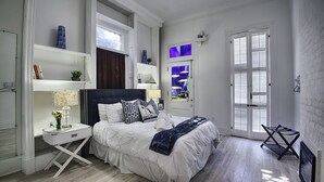 A bedroom with a large bed, white walls, and a wooden floor. There is a window with shutters, a nightstand, and a dresser. - Best Location 7BR Next to FR Quarter! By YouRent (New Orleans)