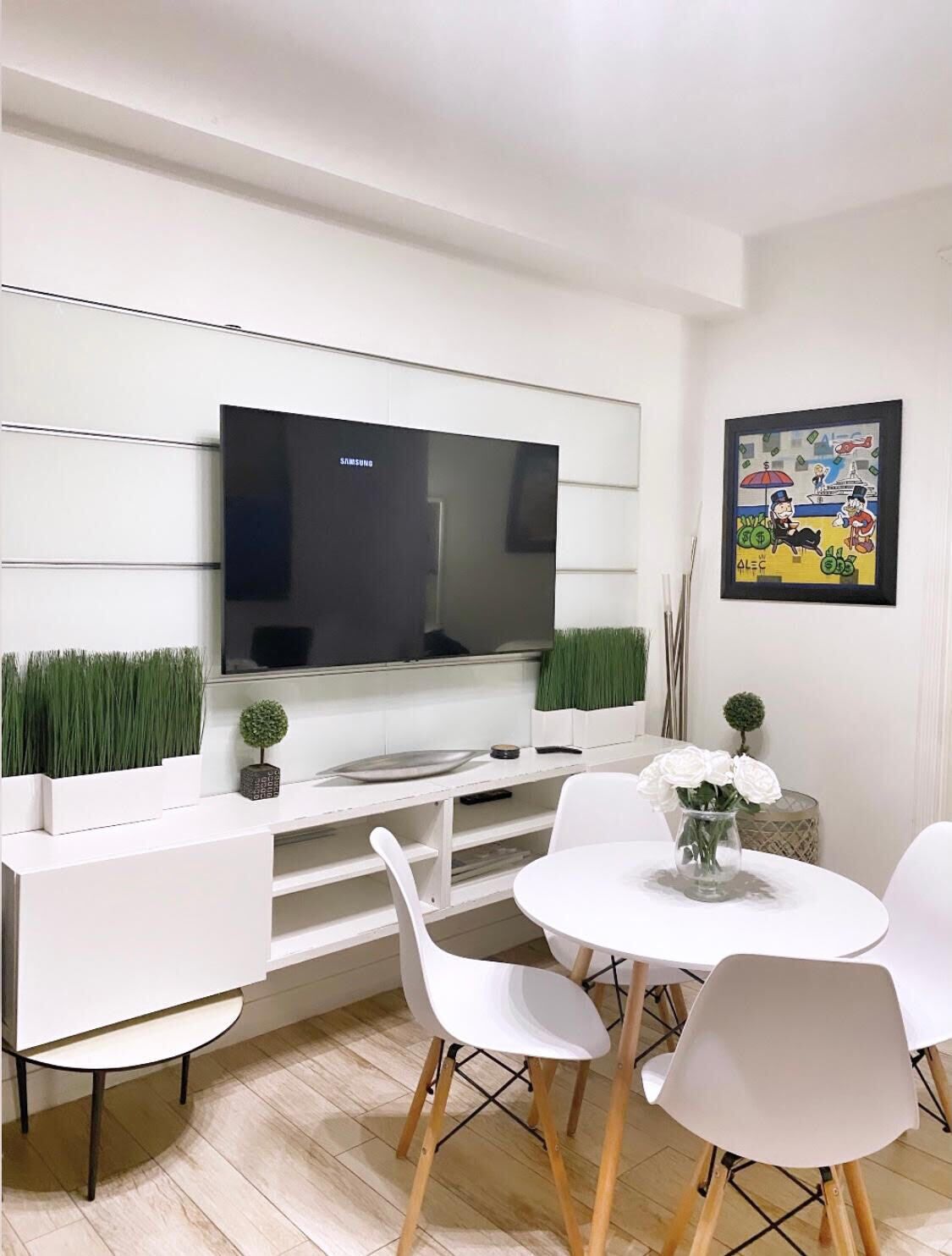 Un salon moderne avec un téléviseur à écran plat fixé à un mur blanc, un coin repas avec des chaises blanches et une table ronde, et une petite plante en pot posée sur la commode.