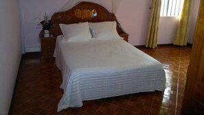 Een netjes opgemaakt bed met een houten hoofdbord, een klein nachtkastje met een lamp en een raam met gordijnen.