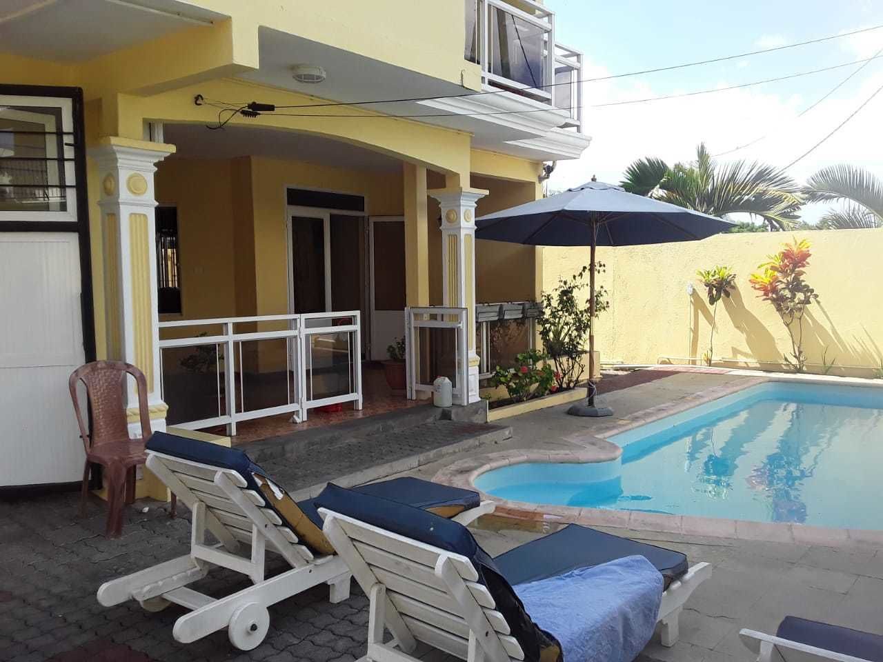 Zona de piscina con sillones de descanso y una sombrilla, un edificio amarillo con balcón y una piscina azul cristalina.