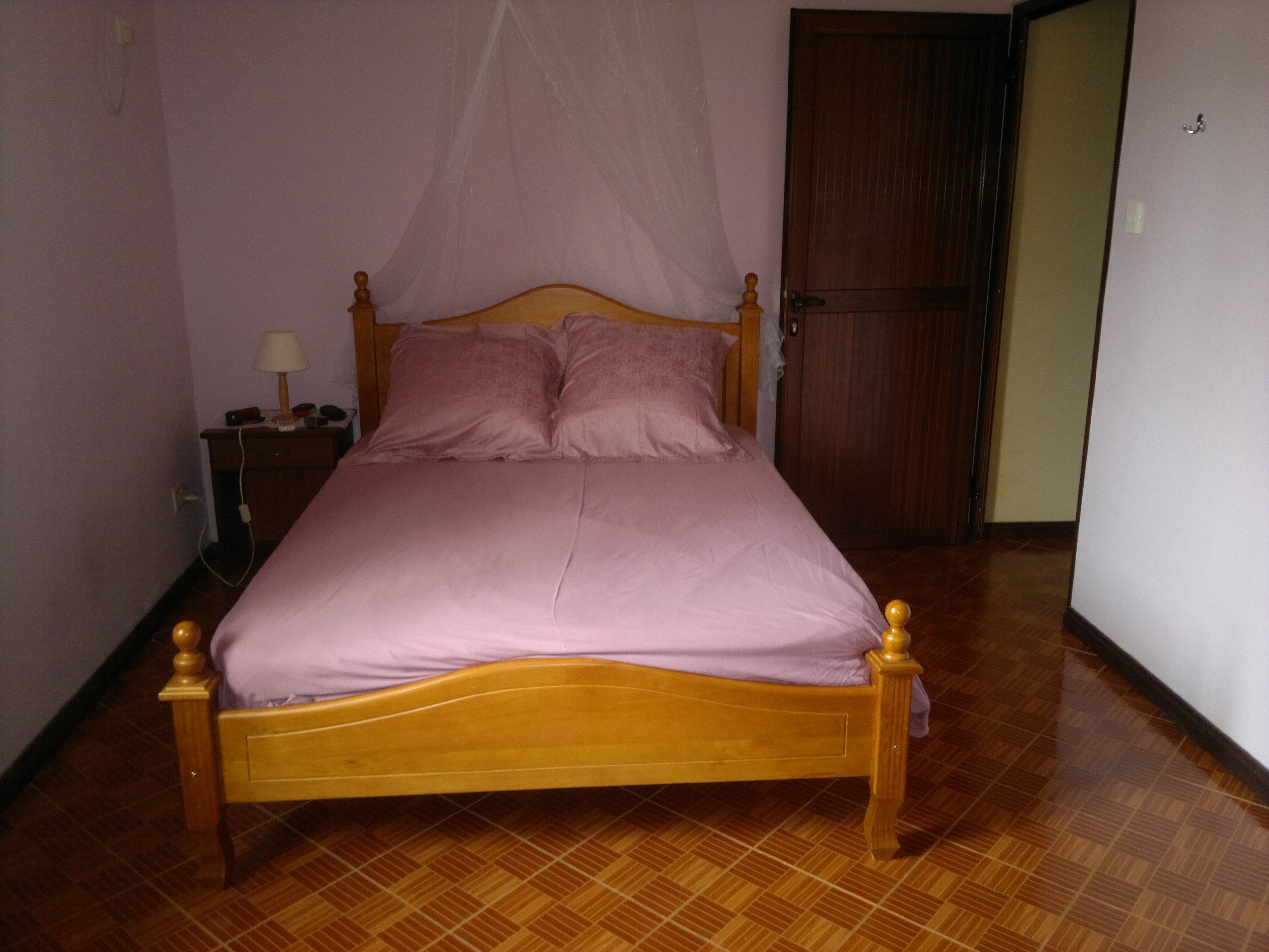 A single bed with a wooden frame and a canopy, a bedside table with a lamp, and a door in the background.