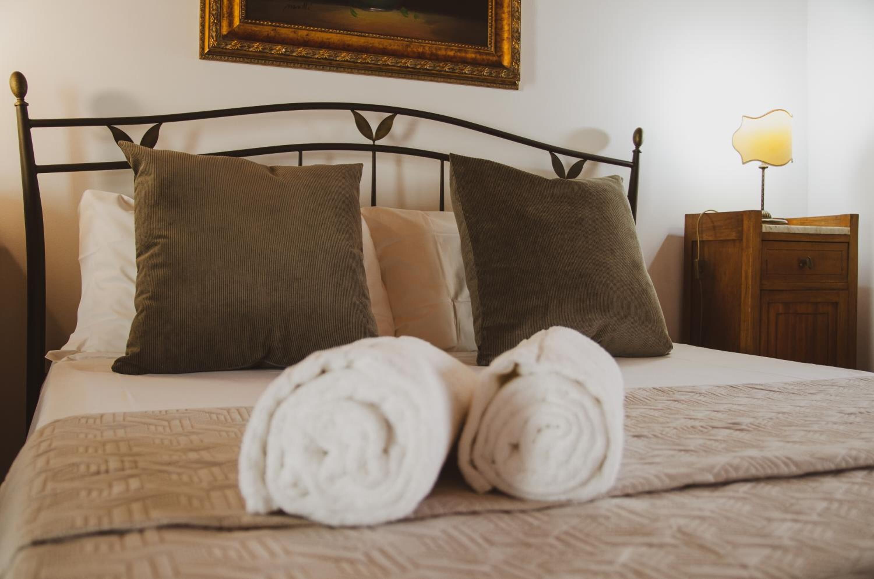 A neatly made bed with a patterned bedspread and rolled towels, a wooden nightstand with a lamp, and a framed picture on the wall.