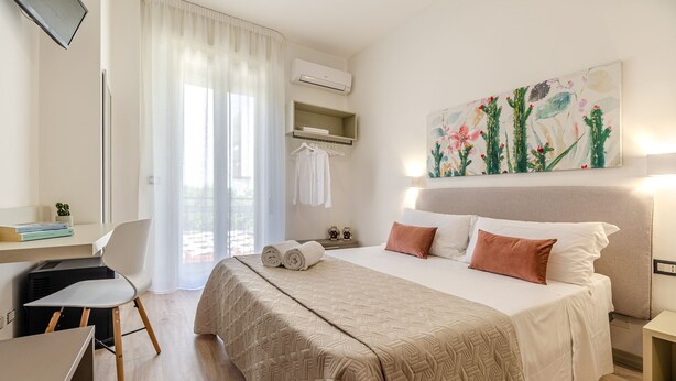 Ein Hotelzimmer mit Bett, Schreibtisch, Stuhl, Fernseher, Klimaanlage, einem Gemälde und einem Vorhangfenster.