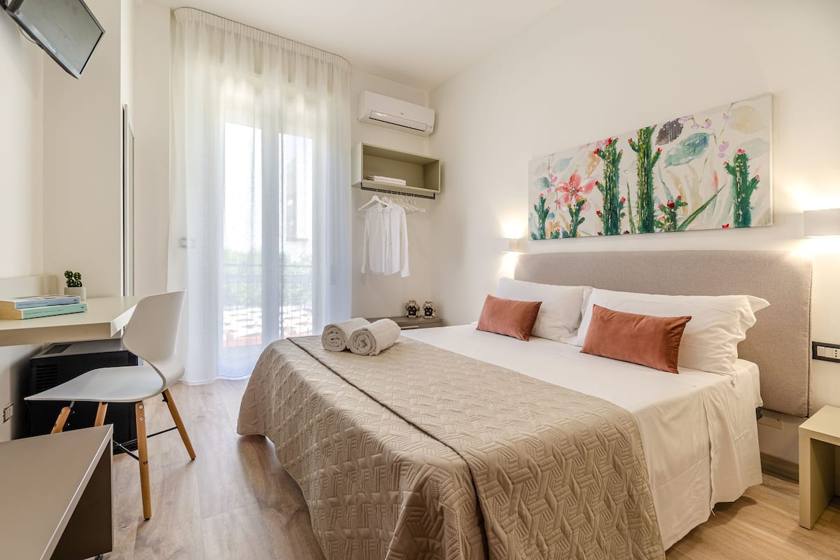 A hotel room with a bed, a desk, a chair, a TV, an air conditioner, a painting, and a window with curtains.