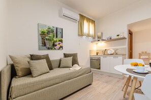 A modern living room with a sofa, a dining table with a cup and oranges, a kitchen area with a stove, and a wall-mounted air conditioner. - Nonna Germina (Roccalumera)