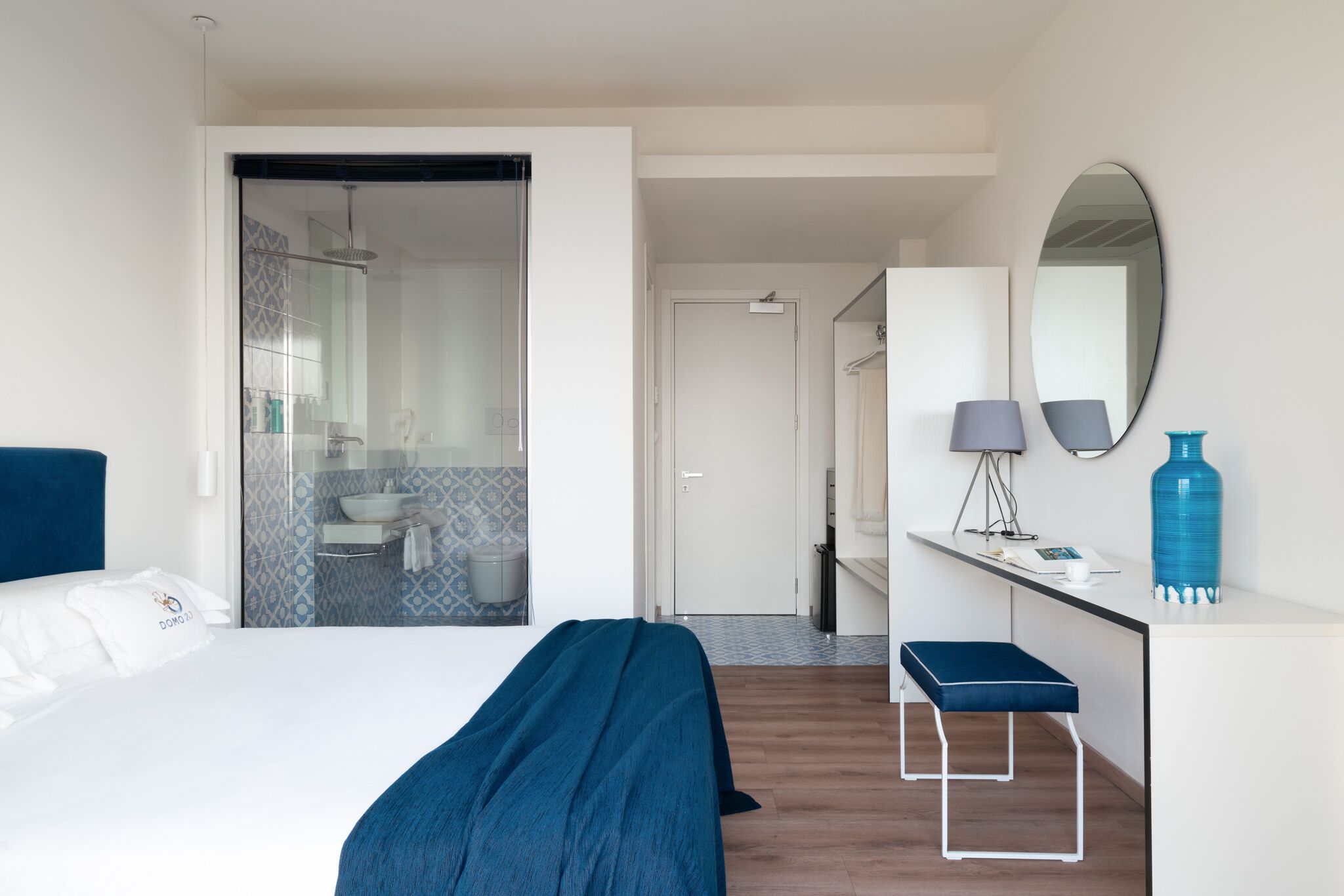 A modern hotel room with a bed, a desk, a mirror, and a bathroom visible through a glass door.