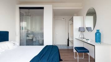 A modern hotel room with a bed, a desk, a mirror, and a bathroom visible through a glass door.