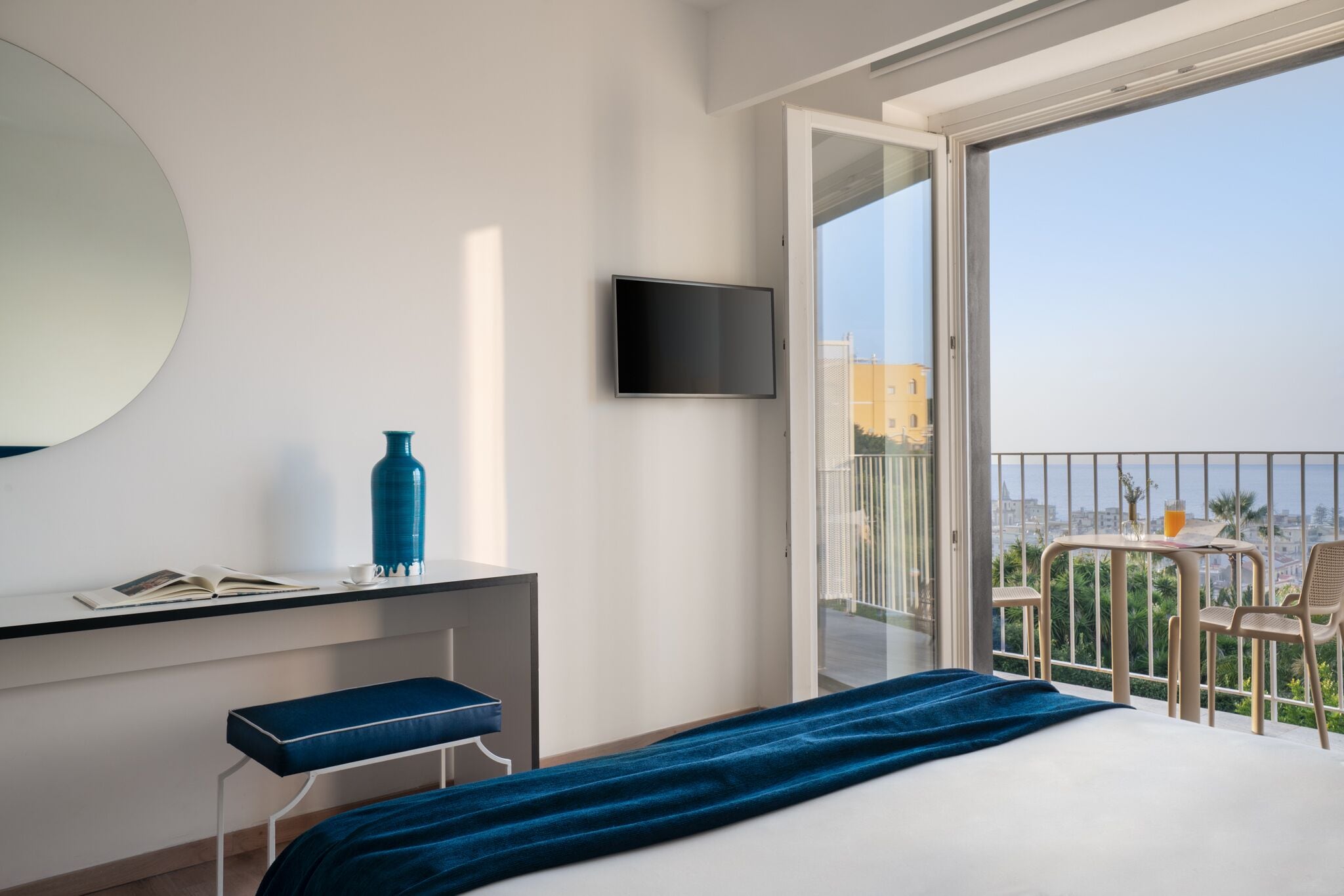 A hotel room with a bed, a desk with a chair, a TV, and a balcony with a table and chairs.