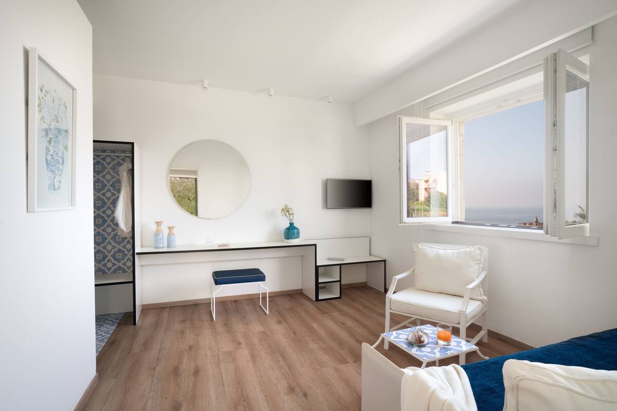 A modern room with a wooden floor, a white desk with a mirror, a TV, a white chair, and a view of the ocean through a large window.