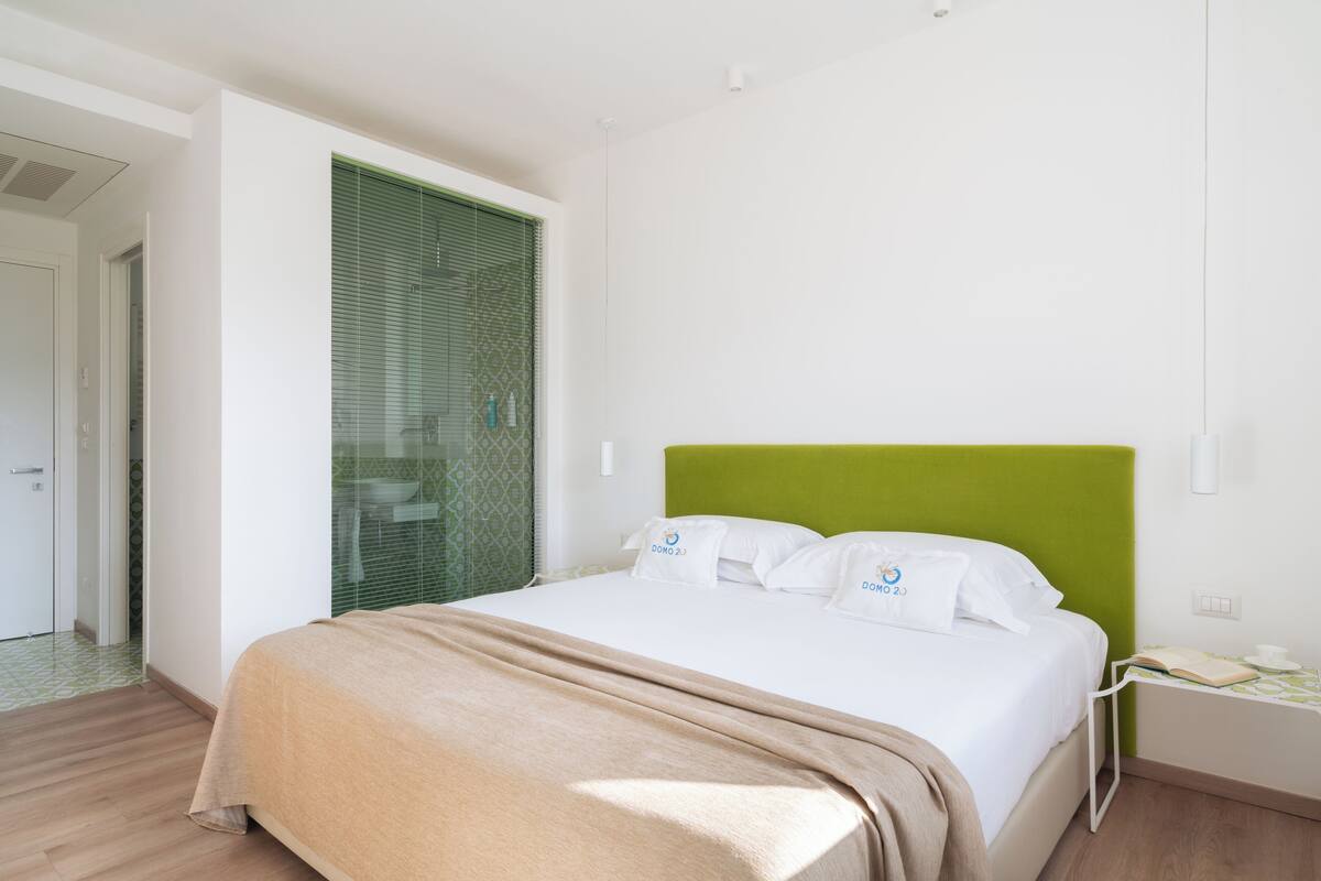 A modern hotel room with a green headboard, white bedding, and a visible bathroom with a green patterned shower curtain.