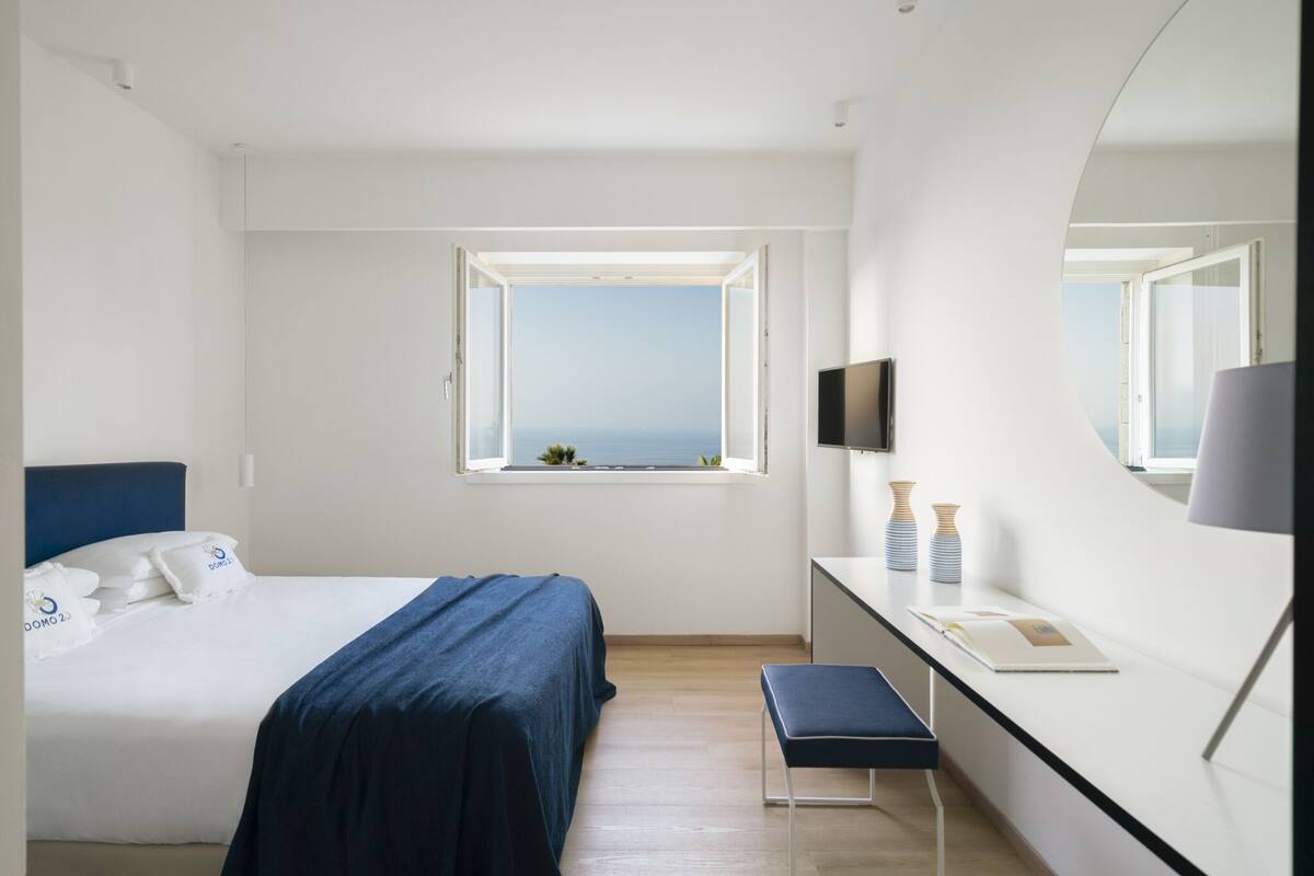 A modern hotel room with a bed, a desk, a bench, a large mirror, and a window with a view of the sea.