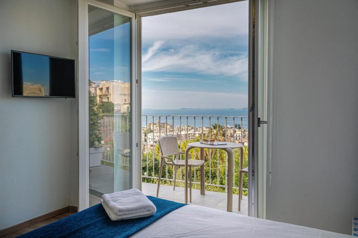 Habitación de hotel con cama, televisión, balcón con mesa y sillas, y vista a la ciudad.