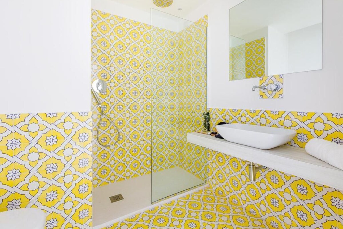 A bathroom with a yellow patterned tile wall, a glass shower enclosure, a white sink, and a mirror.