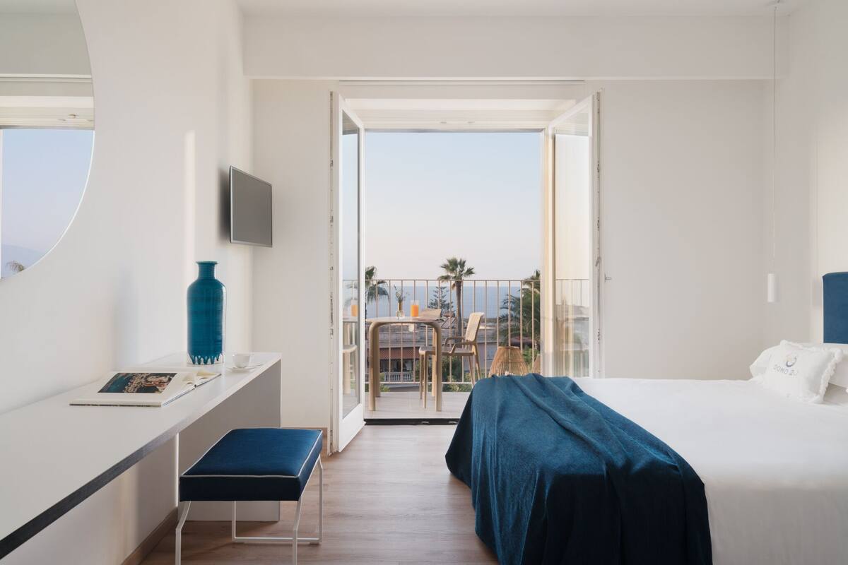 A hotel room with a bed, a desk with a chair, a TV, and a balcony with a view of palm trees and the sea.