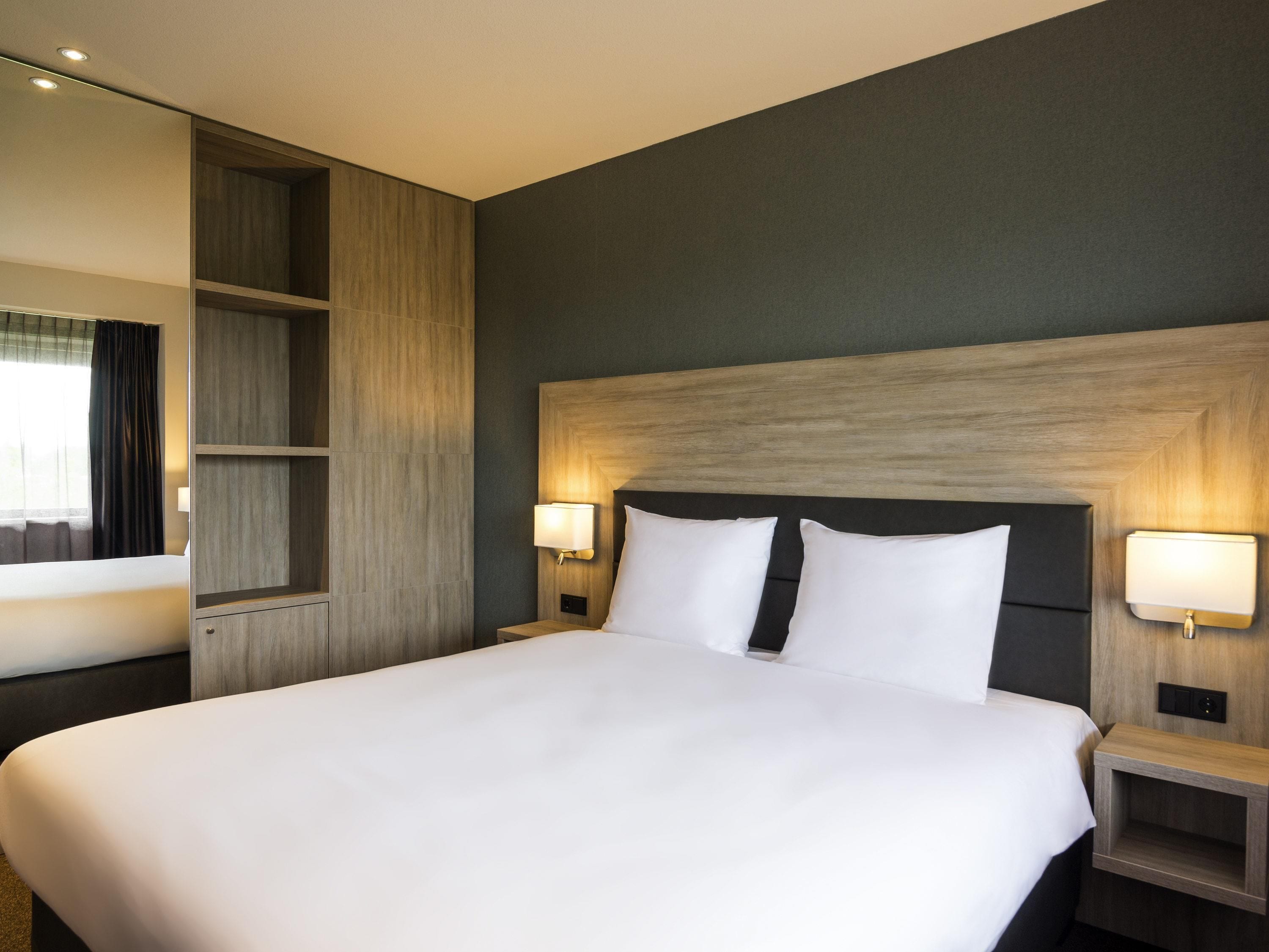 Habitación de hotel con una cama grande, dos mesitas de noche con lámparas, una estantería empotrada y una pared oscura.
