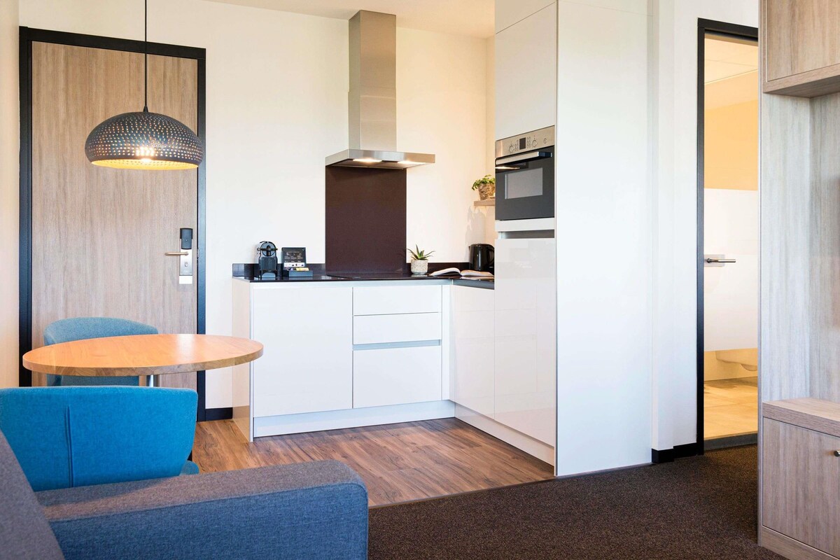 Eine moderne Küche mit weißen Schränken, einem eingebauten Backofen und einer Mikrowelle. Es gibt einen Essbereich mit einem runden Tisch und blauen Stühlen.