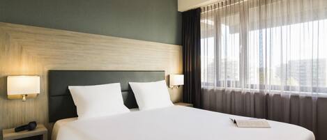 Una habitación de hotel con una cama grande, dos mesitas de noche, un cabecero y una ventana con cortinas.