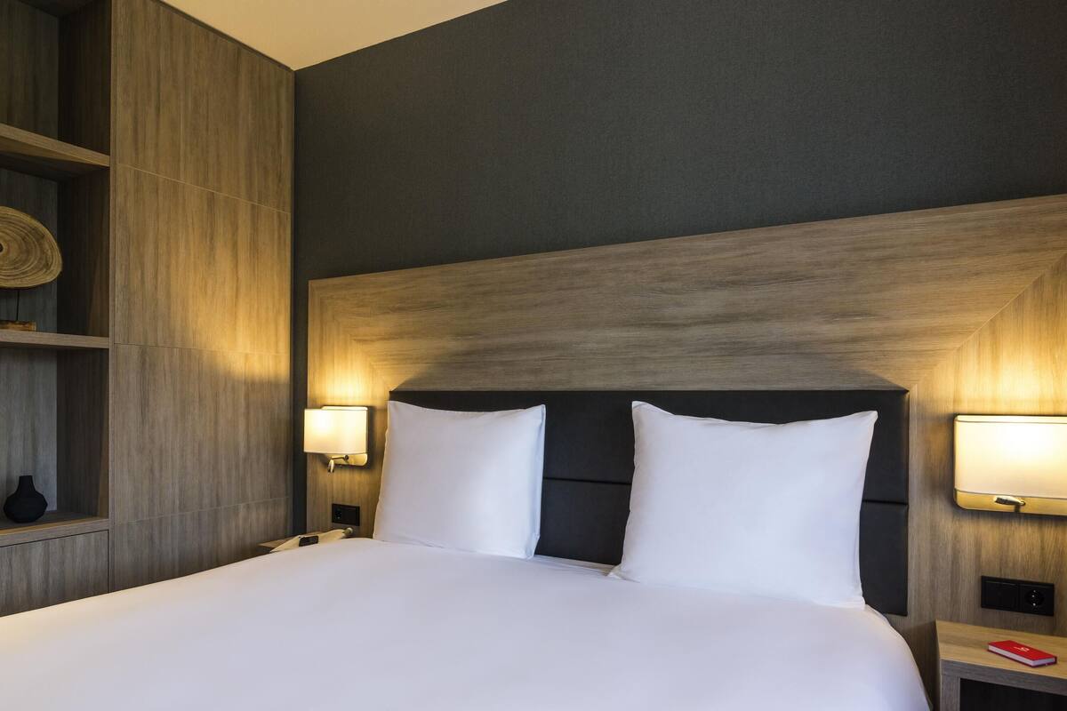 A hotel room with a large bed, two bedside lamps, a wooden headboard, and a small nightstand with a red book.