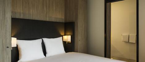 Habitación de hotel con una cama grande, cabecera de madera, dos lámparas de noche y una puerta que da a un baño.