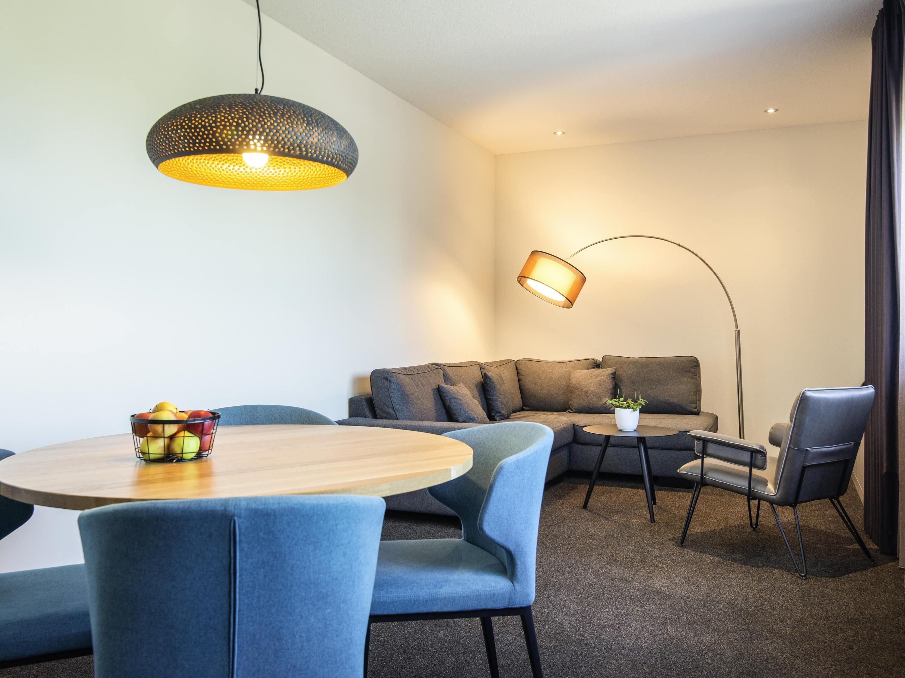 A modern living room with a round wooden table, blue chairs, a grey sofa, a floor lamp, and a hanging light fixture.