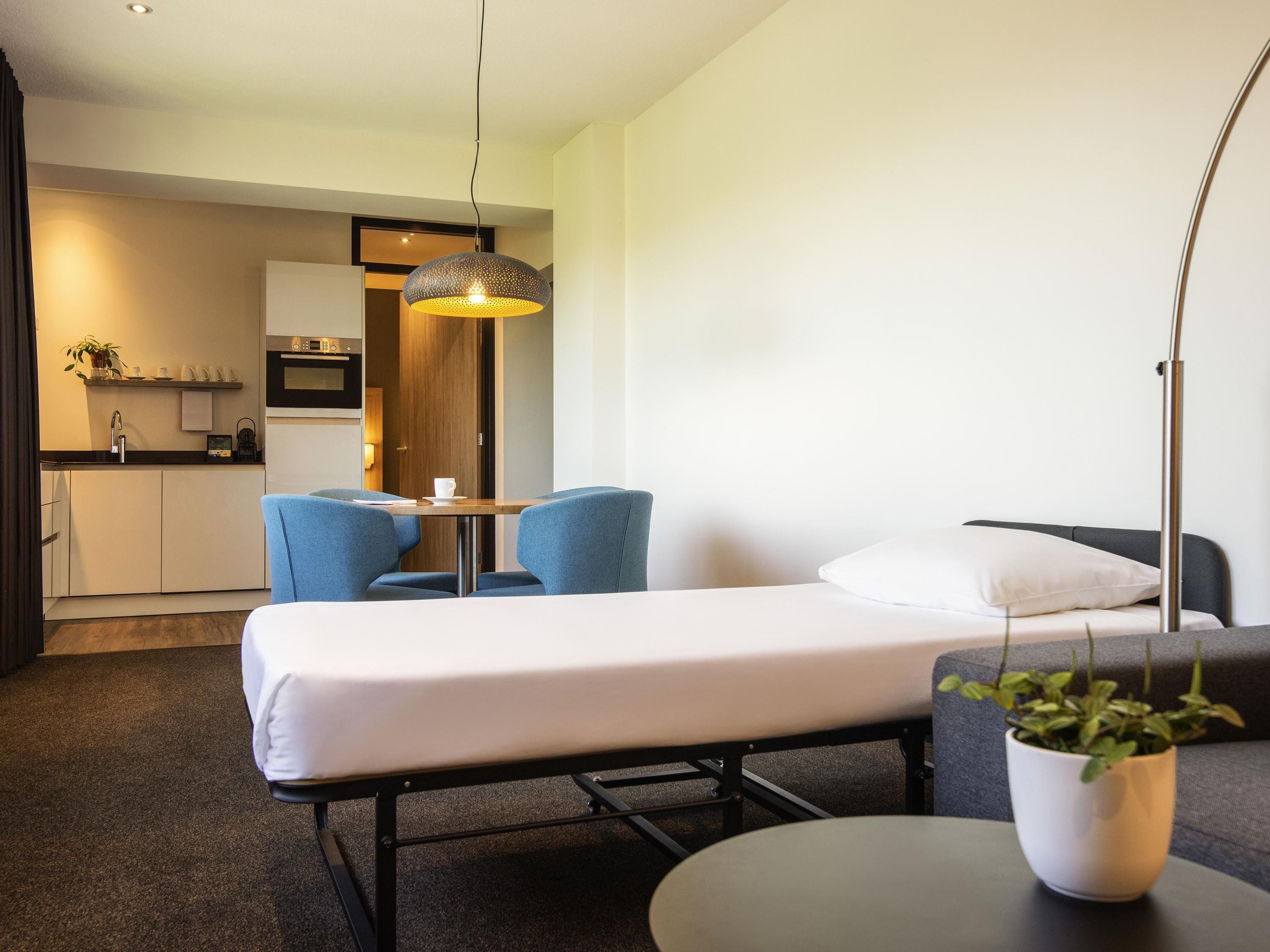 A hotel room with a bed, a small table, two chairs, a potted plant, a floor lamp, and a kitchenette in the background.