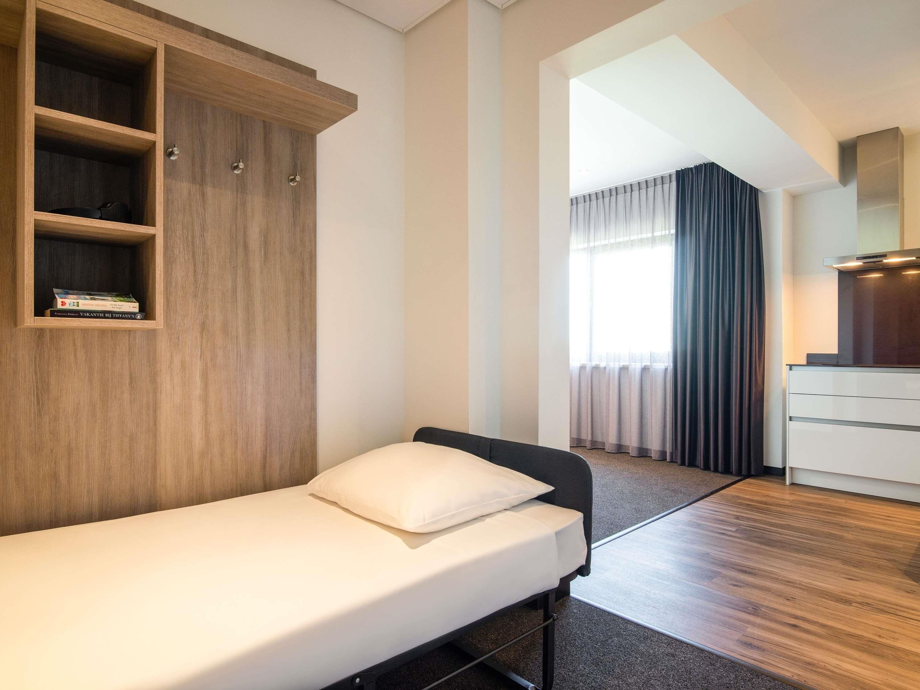A single bed with a white mattress and pillow, a wooden wardrobe with shelves, a white desk with drawers, and a window with curtains.