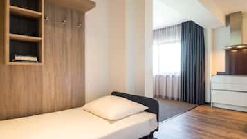 A single bed with a white mattress and pillow, a wooden wardrobe with shelves, a white desk with drawers, and a window with curtains.