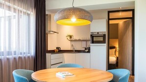 Una habitación de hotel moderna con una mesa redonda de madera, sillas tapizadas en azul, una zona de cocina con microondas y una lámpara colgante grande con textura.