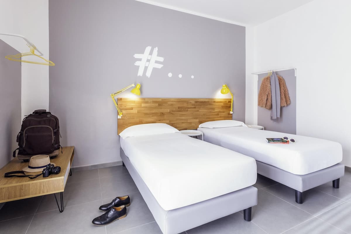 Habitación de hotel moderna con cabecera de madera, dos camas individuales, un pequeño escritorio con silla, perchero y lámpara de pared.