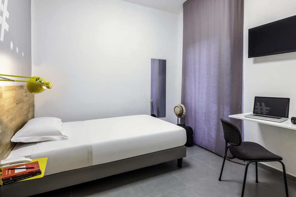 Habitación de hotel moderna con cama, escritorio con una computadora portátil, silla, lámpara y un ventanal con cortinas.