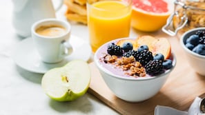 En frukostuppläggning med en skål havregrynsgröt toppad med bär och müsli, ett glas apelsinjuice, ett halvt äpple, en kopp kaffe och en grapefrukt.