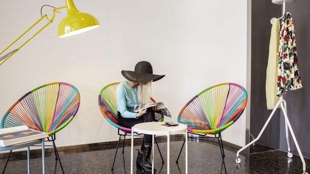 A person seated at a small round table with a magazine, surrounded by colorful chairs, in a room with a plain wall and a floor lamp. - ibis Styles Brindisi (Brindisi)