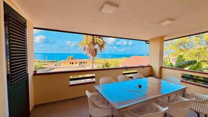 A balcony with a table and chairs, overlooking a coastal view with a palm tree and buildings. - Villa Paglianiti - Your Family Residence (Briatico)