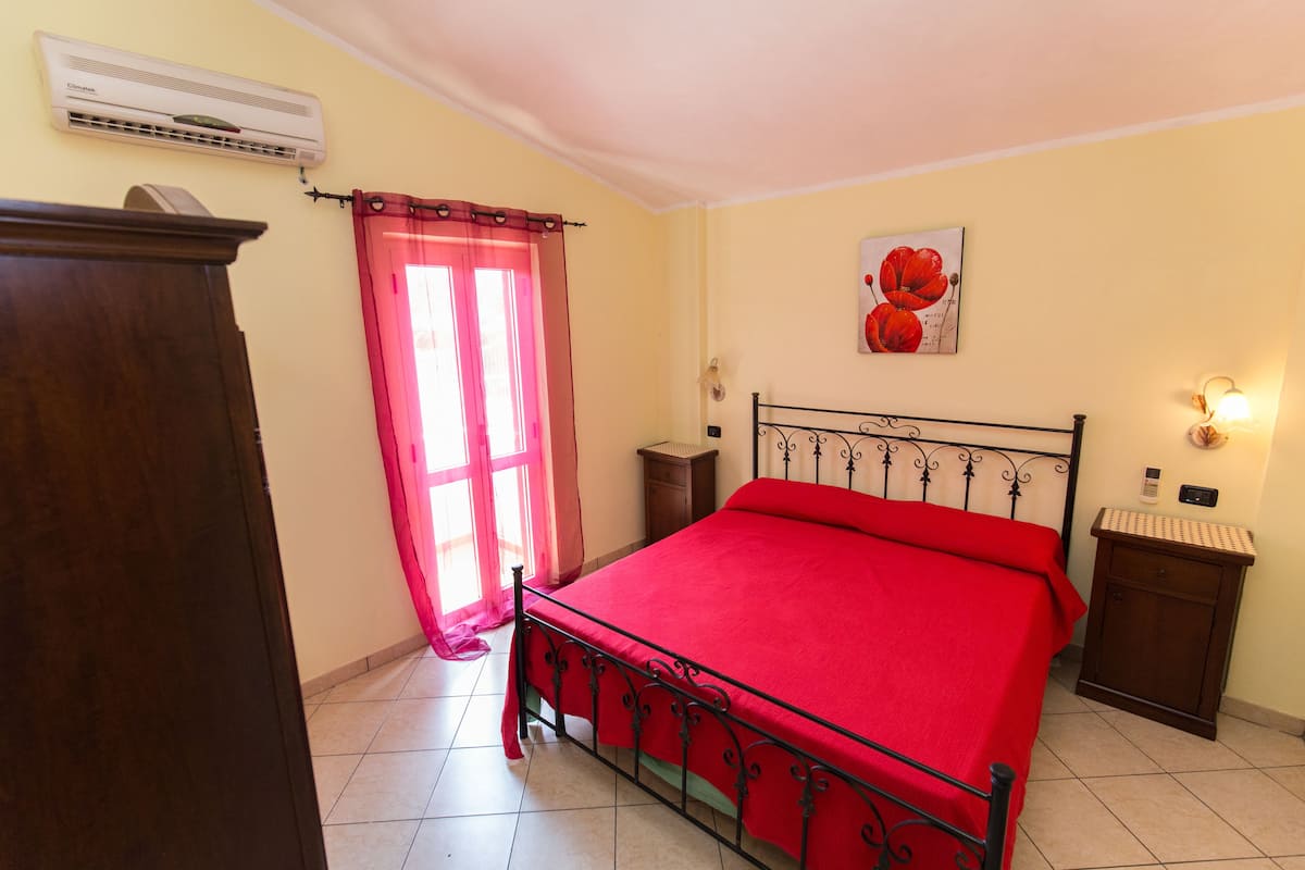 Quarto com cama, mesa de cabeceira, ar condicionado na parede e janela com cortina rosa.