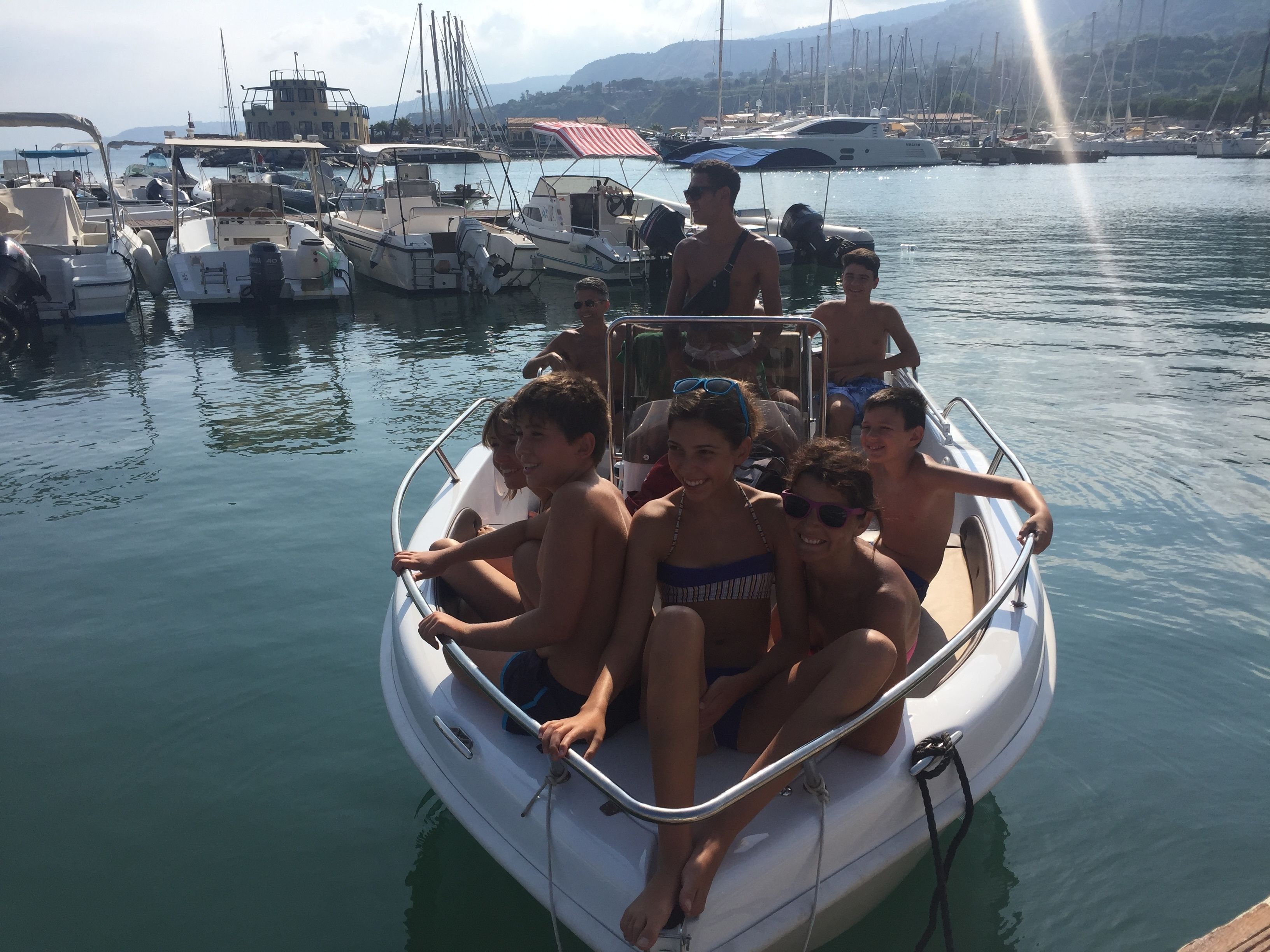 A small boat with several people on board, docked at a marina with other boats and yachts in the background.